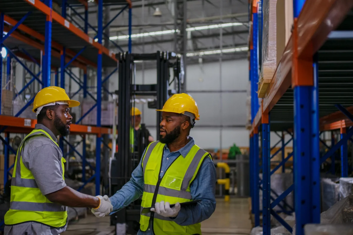 Two workers in hard hats and hi-vis vests shaking hands in a warehouse
