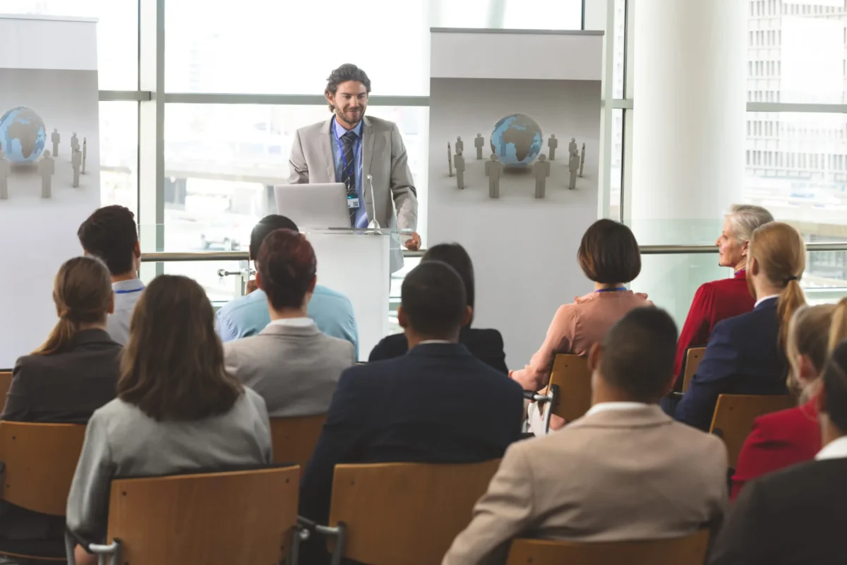 Man presenting at a podium to a seated audience, with globe graphics on banners behind him